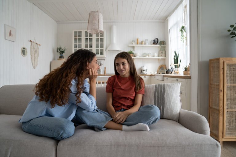 Happy good-natured mother sits on couch at home with teenage girl listening to dreams about future