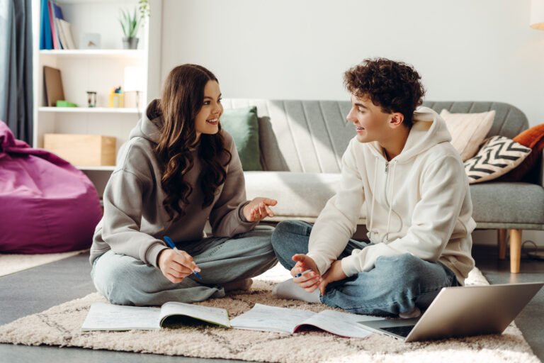 Two happy students collaborating on project at home using laptop and notebooks