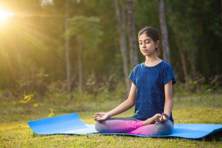 peaceful young indian girl sitting in a padmasana yoga position or lotus pose with her eyes closed in the forest at a sunrise time