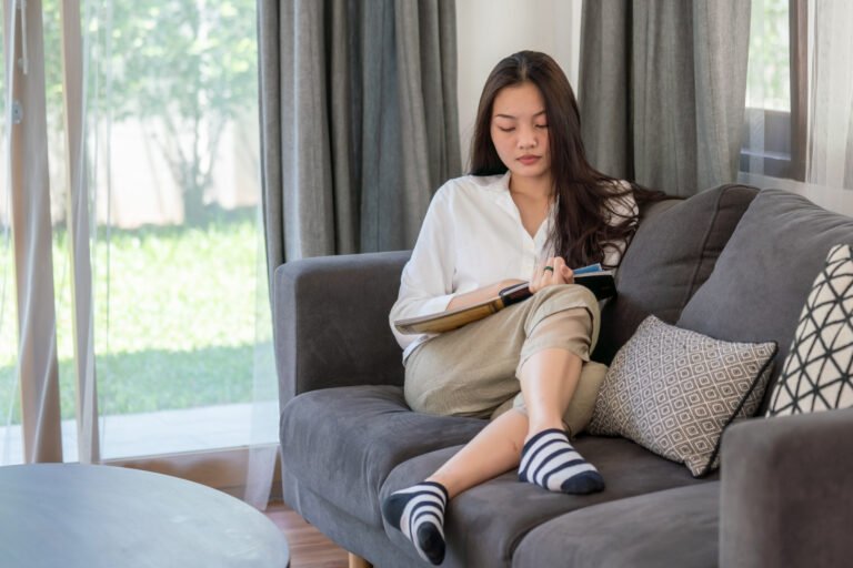 Selective focus of attractive young asian woman lying down on sofa in living room and reading a book alone. Beautiful teenage girl resting and enjoying to read magazine at home in weekend.