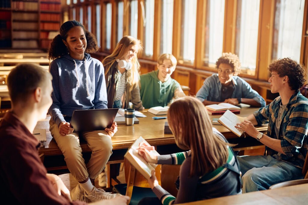 Happy black teenager girl using laptop while studying with friends in library.