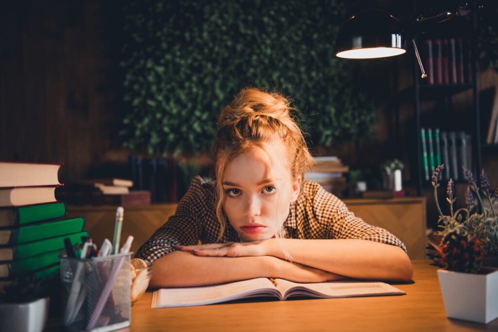 Young woman studying at home with books and lamp light, feeling tired yet determined for exam preparation