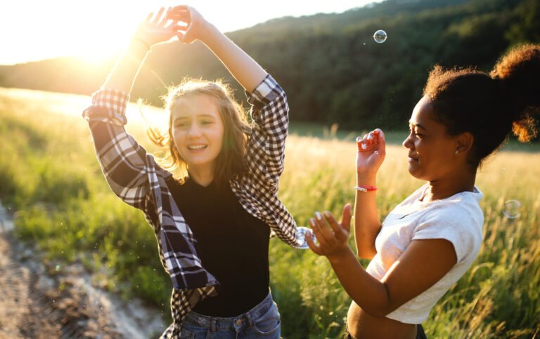 Young teenager girls friends outdoors in nature at sunset, blowing bubbles.