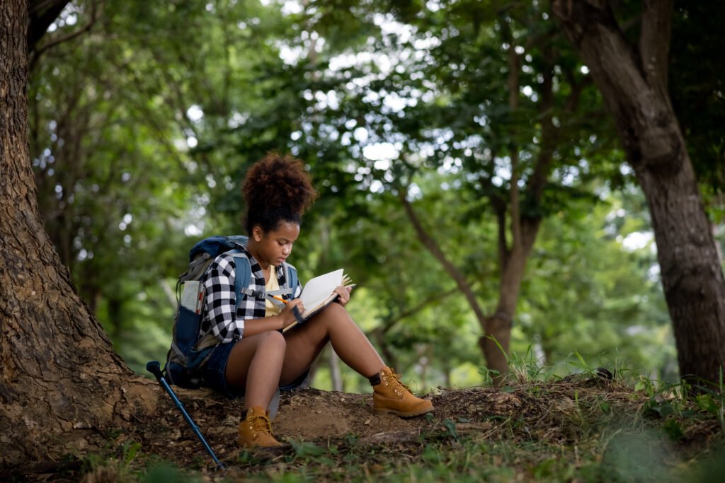 African Ethnicity black skin female teenager hiking backpacker sitting beside a big tree in the natural forest and take note of a notebook explorer trip concept