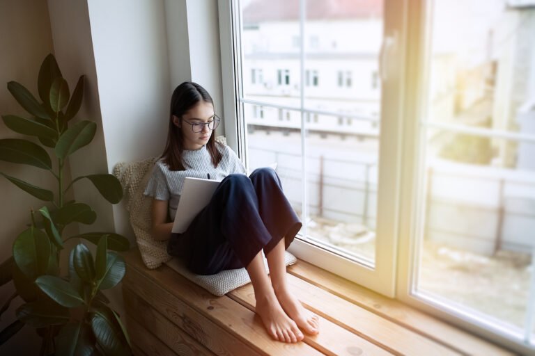 Cute teenager girl reading book at home while sitting on window sill
