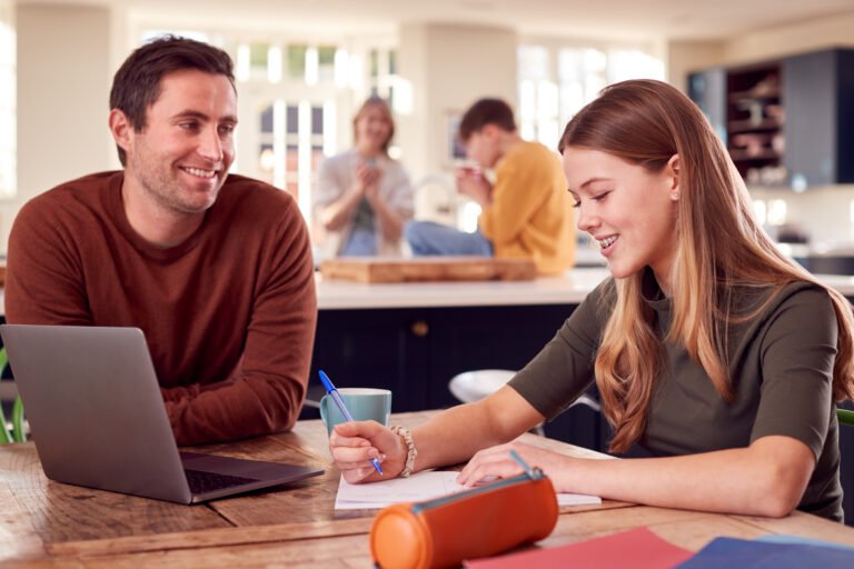 Father Helping Teenage Daughter With Homework Sitting At Kitchen Table At Home Using Laptop
