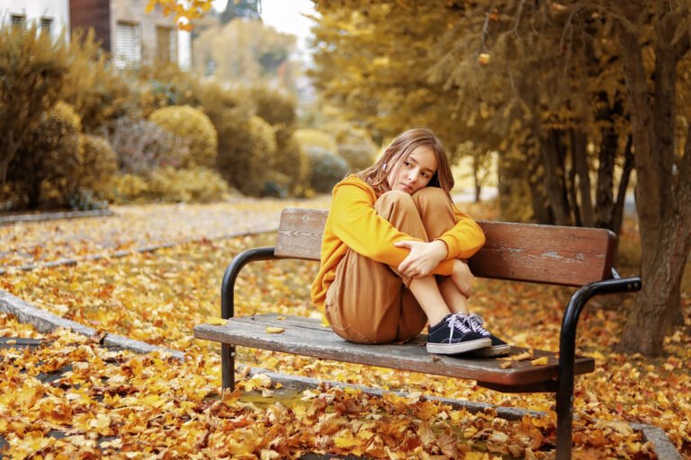 Monochrome autumn. Beautiful teenager girl in yellow hoodie and brown jeans sitting thoughtfully on wooden bench embracing her legs with head on knees in fall park outdoors. First love feelings.