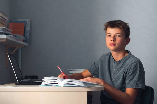 teenager boy doing homework with laptop, writing in copybook
