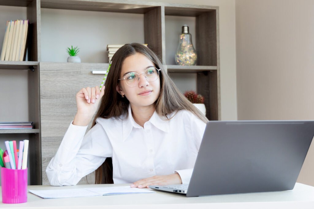 Thoughtful young girl studying online using laptop. A young girl with long hair and glasses sits at a desk, holding a pencil and looking thoughtfully into the distance.