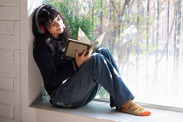 teenage girl with headphones reading a book sitting by a window