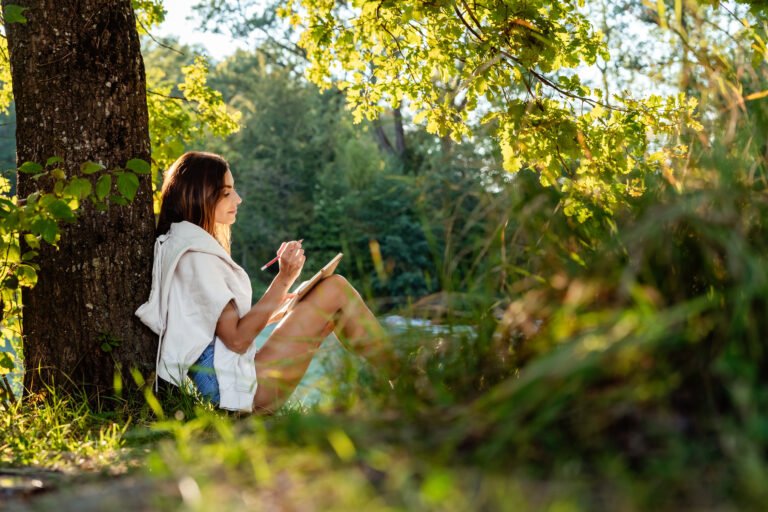 Young attractive girl writing and drawing in notebook sitting under the tree on nature in sunlight. Closeness to nature  inspires creativity. Urban escapism.