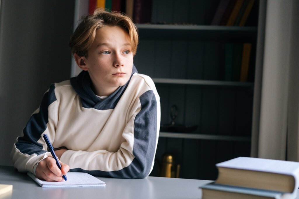 Close-up of pupil boy thoughtful writing in notebook with pen sitting at desk near window.