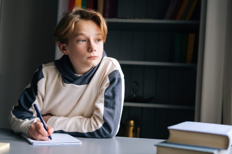 Close-up of pupil boy thoughtful writing in notebook with pen sitting at desk near window.