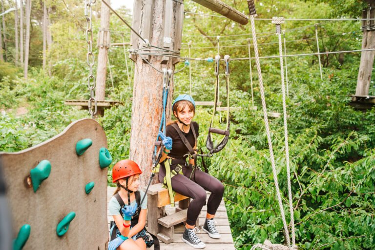 Teenager girls on hinged trail in Adventure extreme rope Park in summer forest.High altitude climbing training of child on adventure track, equipped with safety straps and protective helmet.School girls enjoying activity in a climbing adventure park.
