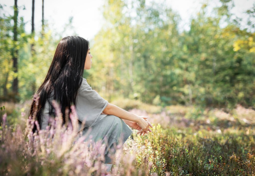 A young woman sitting peacefully in a field surrounded by wildflowers on a sunny day in a serene forest