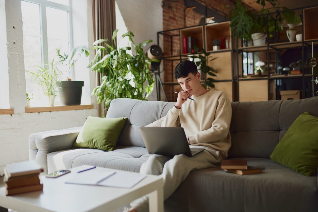 Home Study Session. Young man, student comfortably sitting on grey sofa with laptop at home in modern room and studying online.