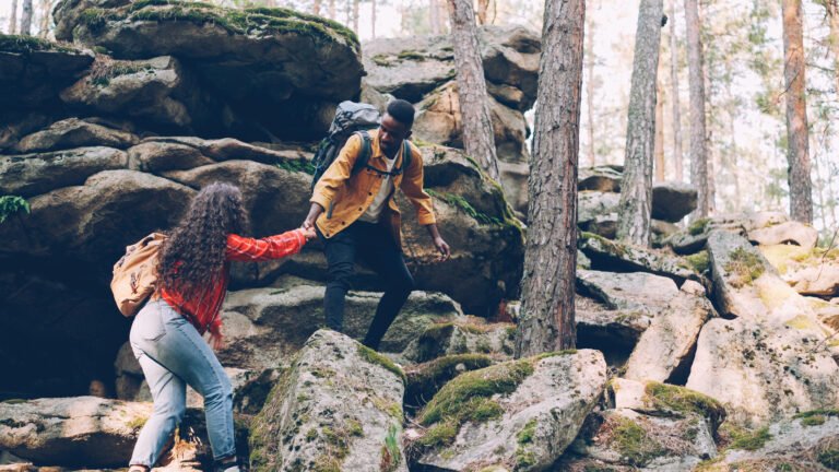 Young multiracial couple is hiking climbing up mountains, African American man is helping woman giving her hand and pulling her up, people are holding backpacks.