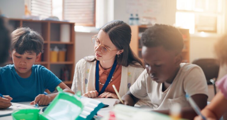 Writing, education and teacher helping kids in classroom for language development, learning or growth. Notebook, advice and woman educator with children students for lesson at elementary school.