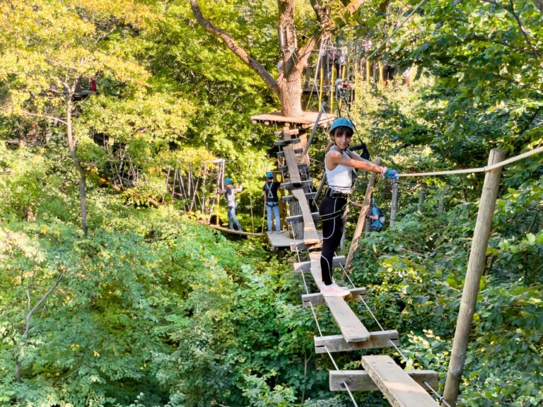 Happy A teenager Girl enjoying activity in a climbing adventure park walks along a rope bridge between trees in safety gear and a helmet on a summer day. Rope park in wood forest.Adventure park close up. Summer fun and sports for adventurous people.