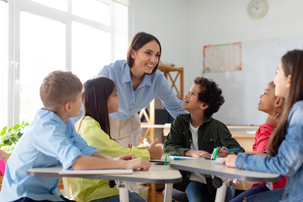 Happy diverse kids sitting at desk in classroom and talking with teacher, studying during lesson, woman explaining subject to pupils