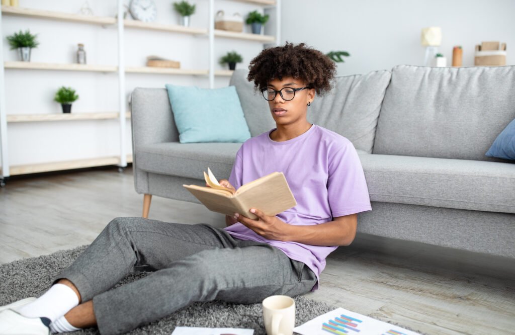 Focused black teenager reading book, sitting on floor, studying from home